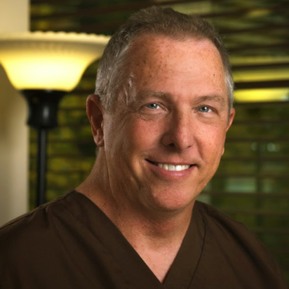 Portrait of Dr. Steven E. Lynn, DDS, a Bloomington implant dentist, smiling and wearing dental scrubs with a lamp and blinds in the background.