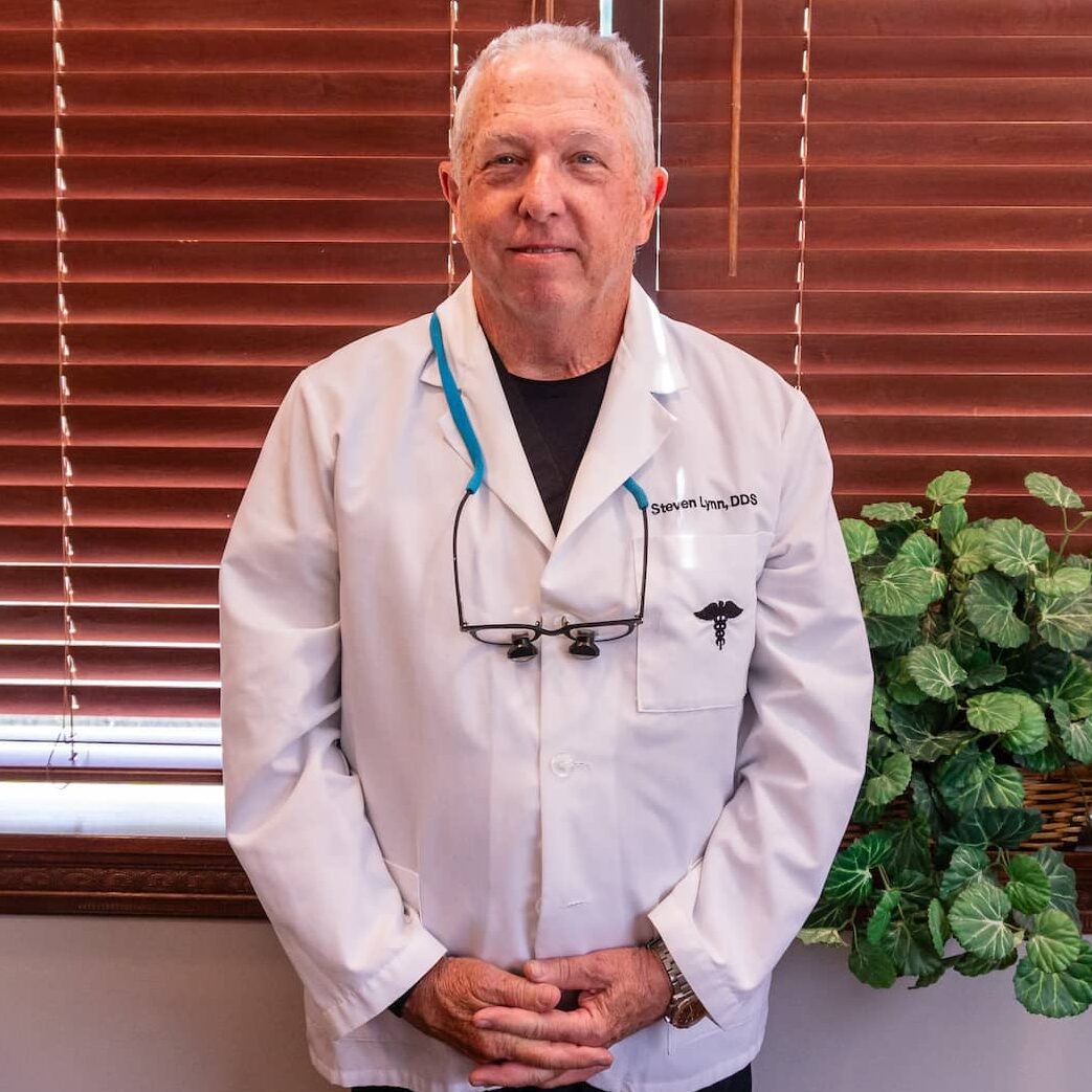 Portrait of Dr. Steven E. Lynn, DDS, a Bloomington implant dentist, smiling and wearing dental scrubs with a lamp and blinds in the background.