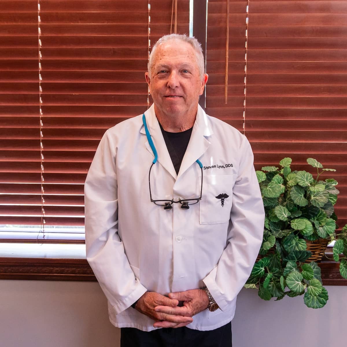 Portrait of Dr. Steven E. Lynn, DDS, a Bloomington implant dentist, smiling and wearing dental scrubs with a lamp and blinds in the background.