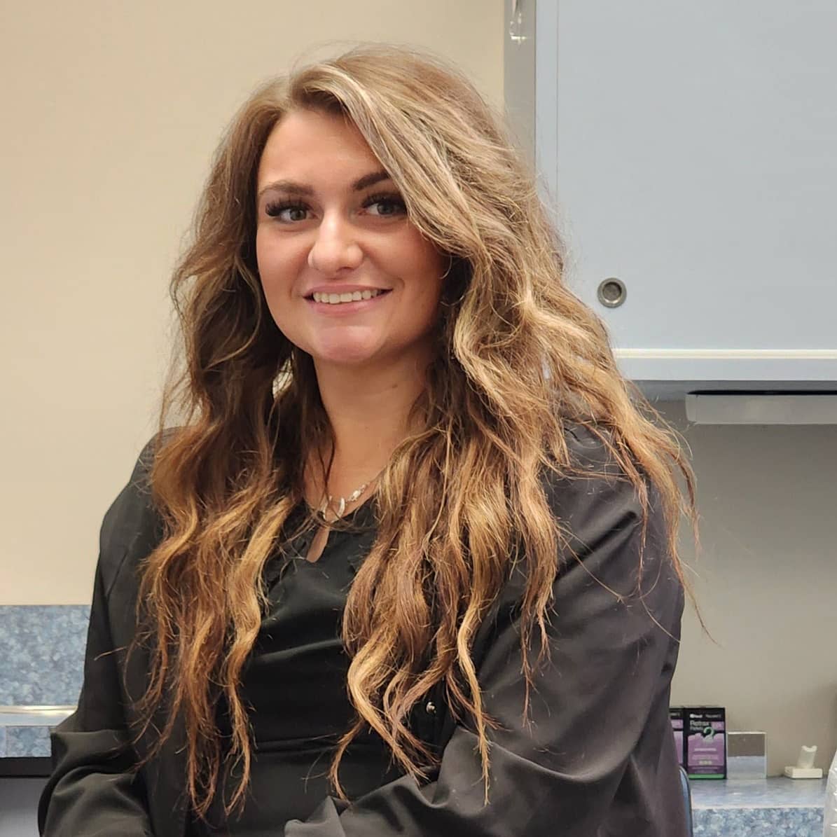 A smiling chair-side dental assistant with long wavy hair, wearing black scrubs, in a dental office.