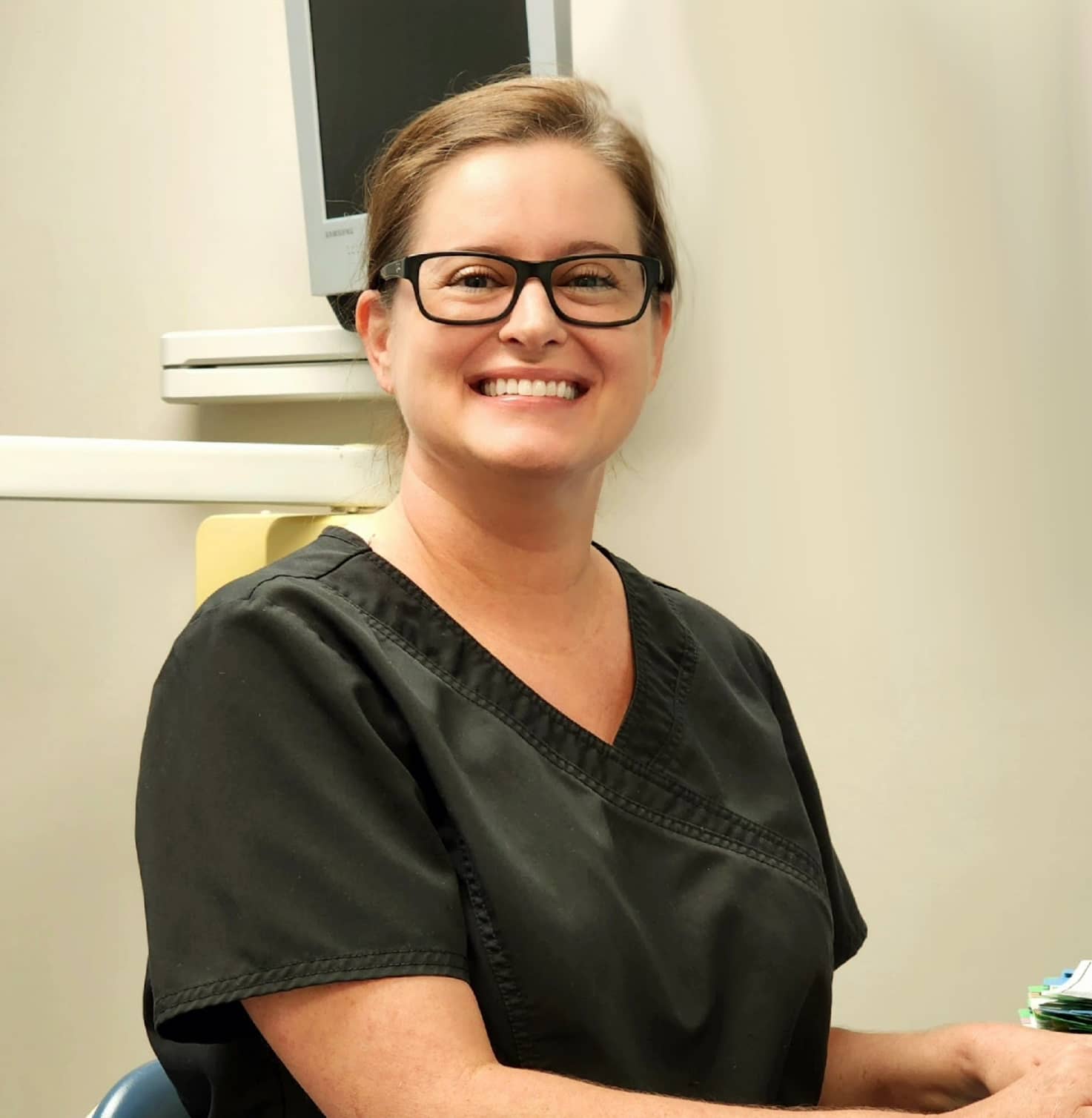 A smiling dental hygienist in black scrubs working at a computer station in a dental office.